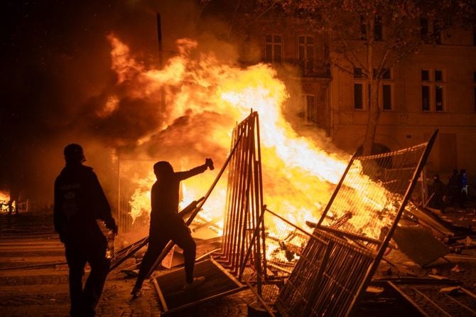 2026年10月17日、フランス・パリのオスマン大通りで、金属製のバリケードの背後で燃え盛る大きな火のそばに立つ2人の若者の夜間のショット。倒れたバリケードや瓦礫の中に立つ一人の男性が、腕を上げている。