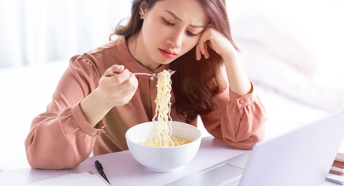 パソコンの前でインスタントヌードルを渋々食べている女性