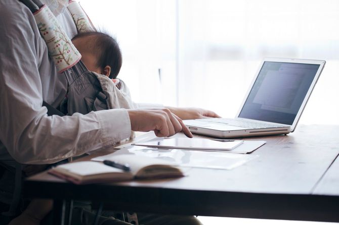 抱っこひもで乳児を抱きながらタブレット端末で仕事をする父親