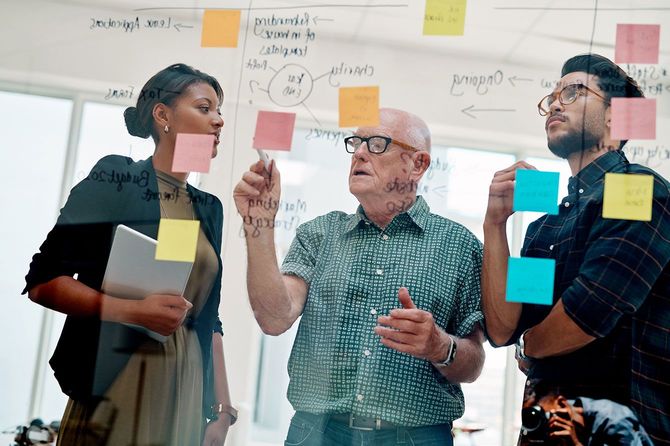 オフィスのガラスボードにメモを書いて若者に説明をする高齢の男性