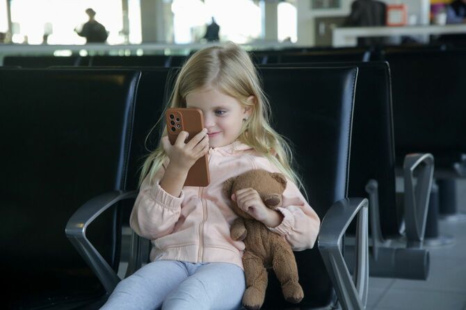 空港の出発ホールに座ってテディベアを持ち、携帯電話で満足そうに遊ぶ少女