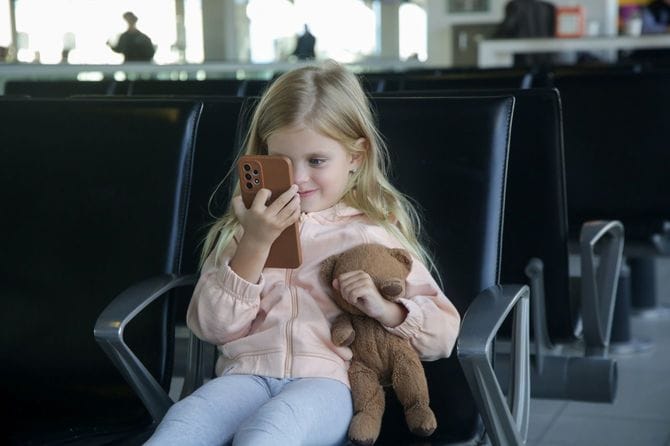 空港の出発ホールに座ってテディベアを持ち、携帯電話で満足そうに遊ぶ少女
