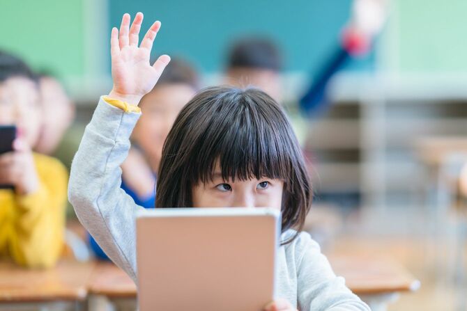 デジタルタブレットを持って教室で手を上げる子ども