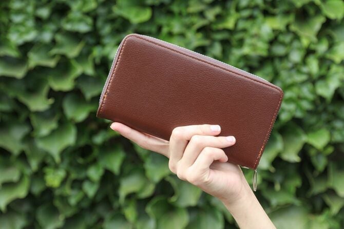 緑の葉の壁の背景に茶色の革の財布を持つ女性の手
