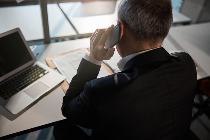 オフィスでノートパソコン、書類を広げたデスクで電話をかけている中年男性
