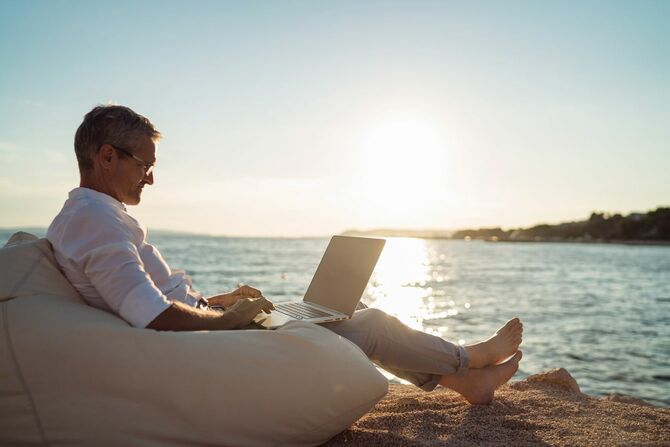 夕暮れ時のビーチでソファに横たわりながらノートパソコンで作業するシニア男性