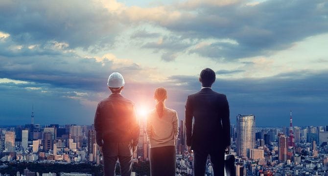 太陽に照らされた高層ビルが立ち並ぶ街を背景にした3人のビジネスパーソンの後ろ姿