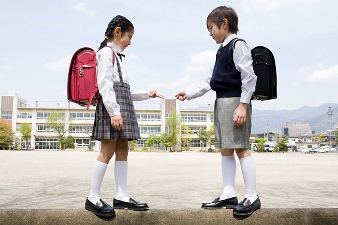 制服を着てランドセルを背負った二人の子供が小学校のグラウンドの塀の上でじゃんけんをしている