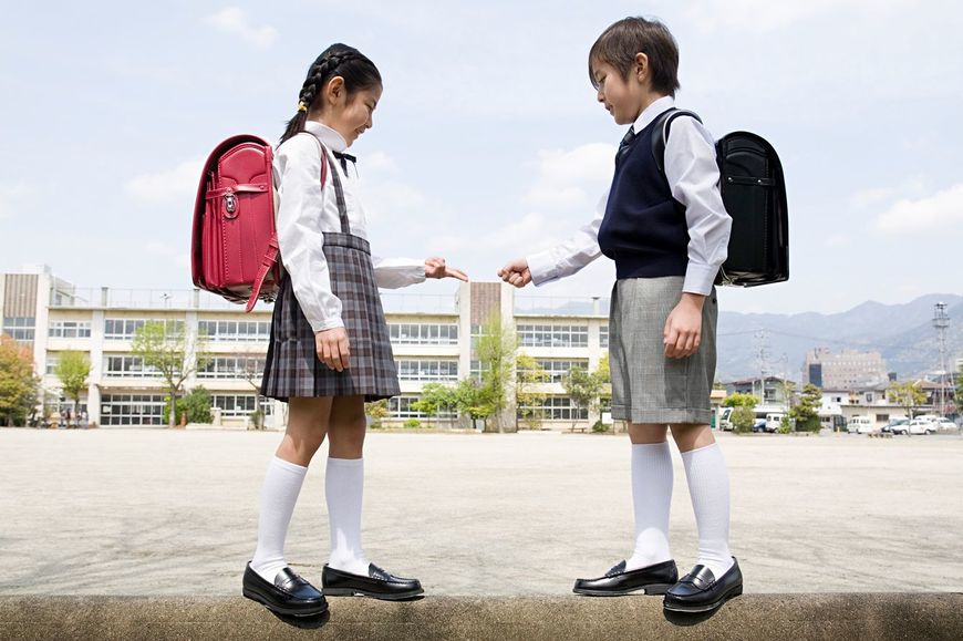 制服を着てランドセルを背負った二人の子供が小学校のグラウンドの塀の上でじゃんけんをしている