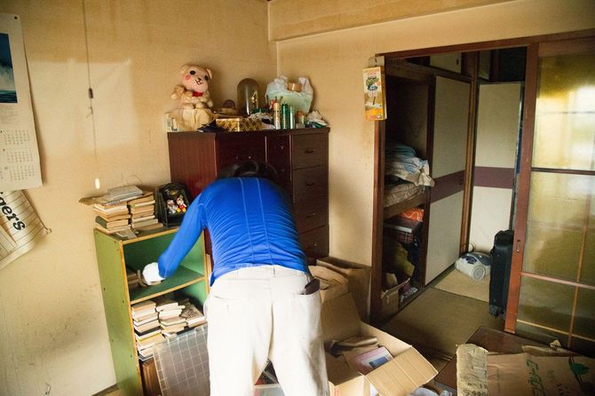 段ボールや遺品が散乱する部屋で本棚を片付ける作業員