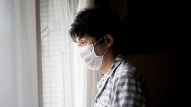 マスクを着用した男性が暗い部屋から外を見ている