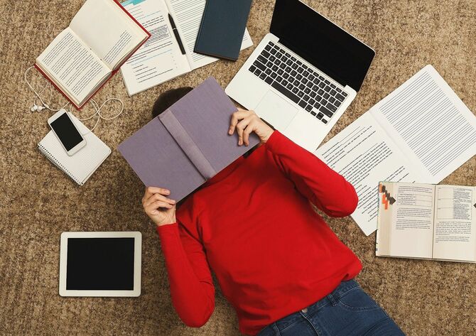 書籍、ガジェットに囲まれて床に横たわり疲れている学生の女の子
