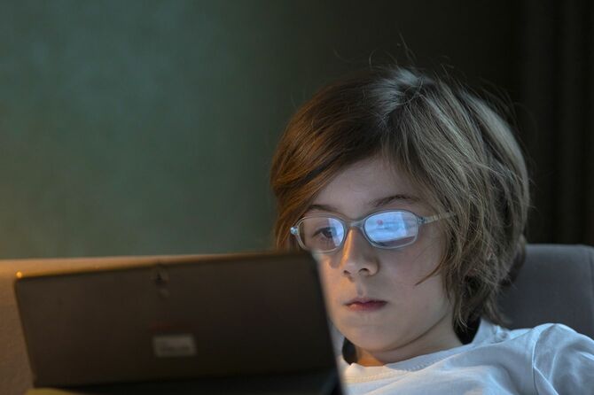 暗い部屋でタブレットを見つめる少年の眼鏡に光が映り込んでいる