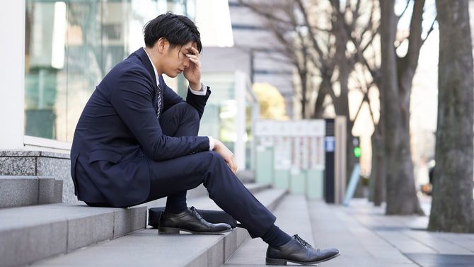 路上の階段に座り込んで頭を押さえる日本人ビジネスマン