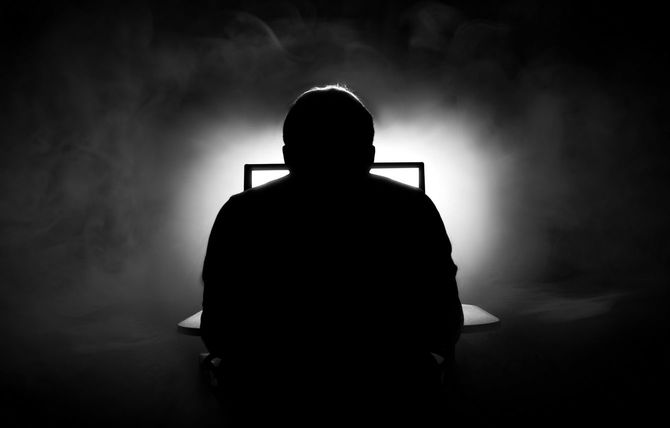 暗い部屋でこうこうと光るパソコンに向かう男性の後ろ姿