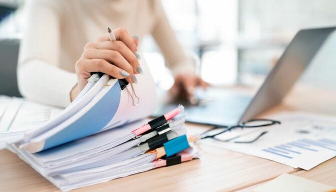 ラップトップで作業しながら、整理された書類の束をペンで持ちながらマルチタスクをしている女性