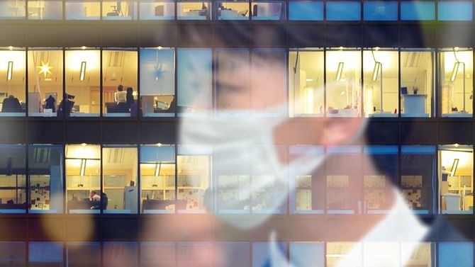 マスクを着けた男性が明かりがつくオフィスビルのガラスに写っている