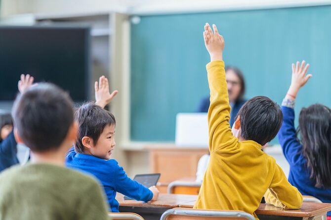 小学校の建物の教室で手を挙げて質問したり答えたりしている男女の小学生のグループの背面図。