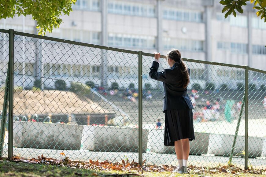 背丈ほどのフェンスに手をかけ、フェンス越しに学校を眺める女子生徒