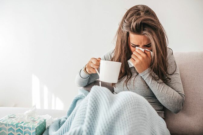 風邪をひいて、ベッドの上で片手にマグカップを持ちながら鼻をかむ女性