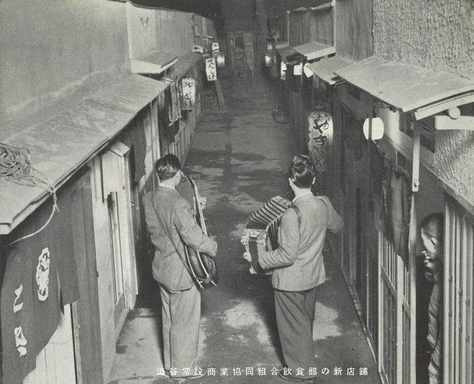 平屋に挟まれた通りに二人の男性が立つモノクロ写真