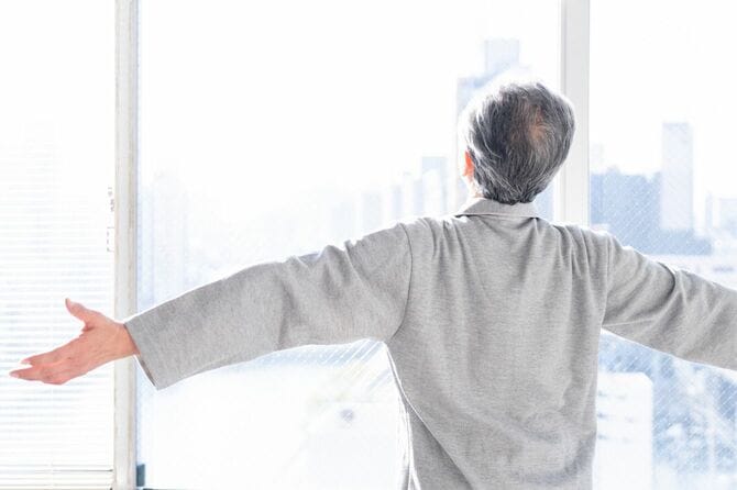 朝に窓の前で腕を広げてリラックスする高齢男性