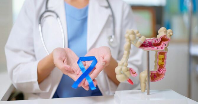 大腸の模型と並んで、大腸がん疾患啓発活動「ブルーリボンキャンペーン」のブルーリボンを差し出す医師の手元
