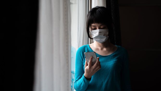 窓の近くでスマホをチェックする、マスクを着けた女性