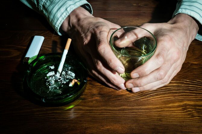 灰皿に乗るタバコとアルコールの入ったグラスを持つ男性