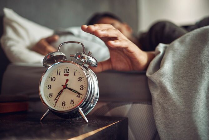 ベッドで目を覚ました後、目覚まし時計に手を伸ばす若い男