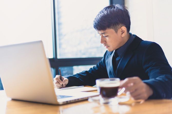 テーブルに座ってラップトップの前でコーヒーを片手にメモを取るビジネスマン