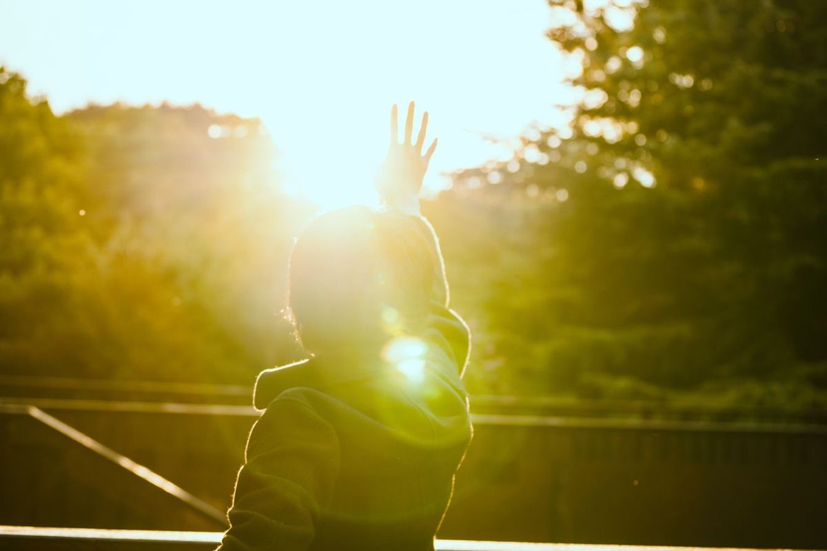 太陽に向かって手を伸ばす男性を背中から見る写真
