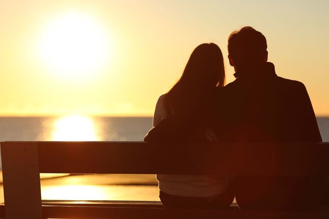 海に沈みゆく夕日をベンチに腰掛け見つめるカップル