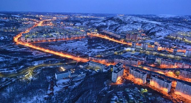 晴れ間がのぞく、わずかに曇った空の下にあるムルマンスクの写真。手前の草原の奥には、ソ連時代の都市計画に典型的な灰色の建物群が並んでいる