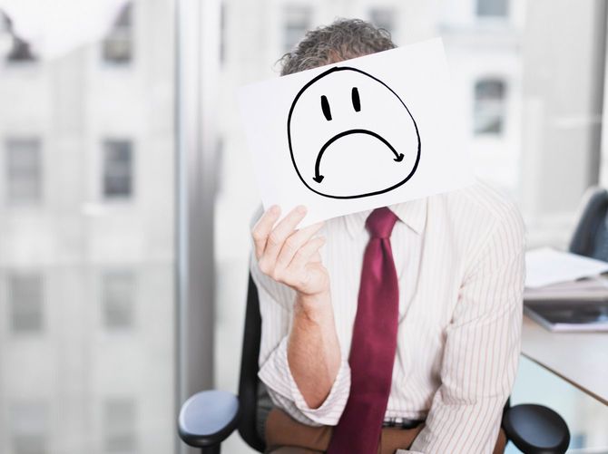 悲しげな顔の描かれた紙を顔の前に掲げるビジネスマン