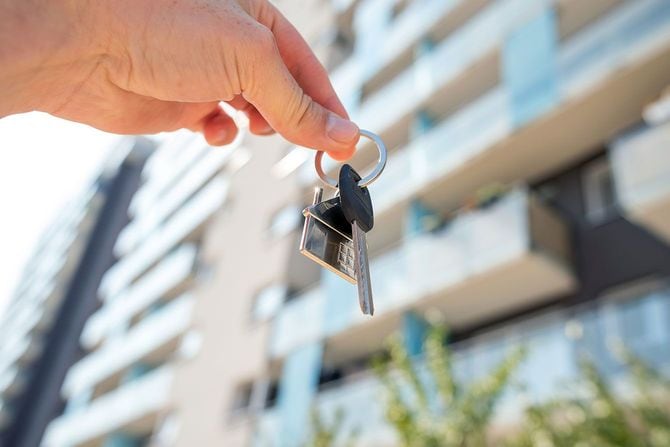 マンションを背景に、家の形のキーホルダーがついた鍵を持つ手