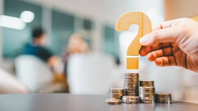 積み上げられた硬貨の上に疑問符のオブジェを乗せるビジネスマンの手