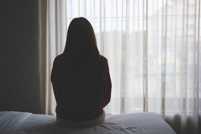電気のついていない部屋でベッドに腰かけ外を見つめている女性
