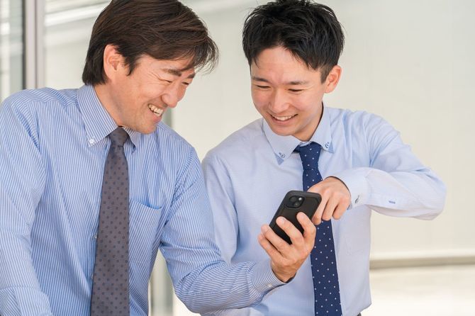男性ビジネスマンがオフィスの休憩室のベンチでスマホを見ながら笑顔で話している