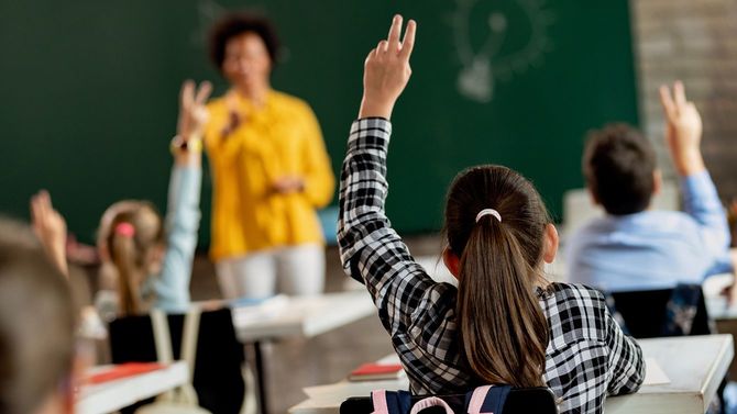 教室での質問に答えるために腕を上げる女子高生の背面図。