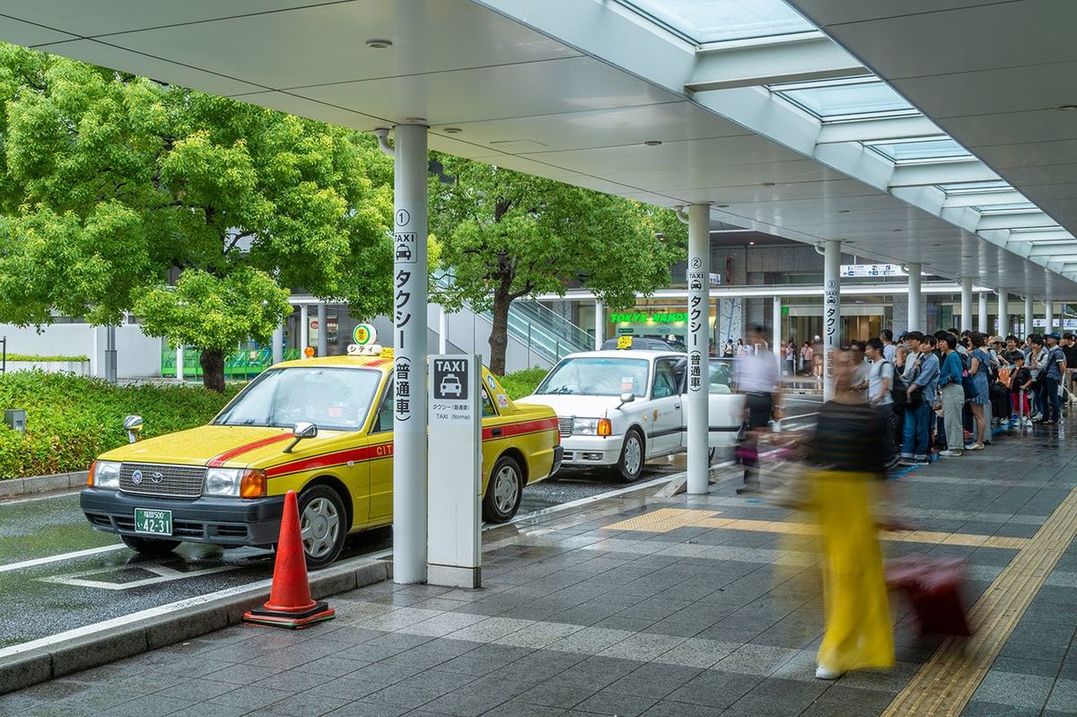 福岡の博多駅ビルの外の歩道でタクシーを拾うために列に並ぶ人々