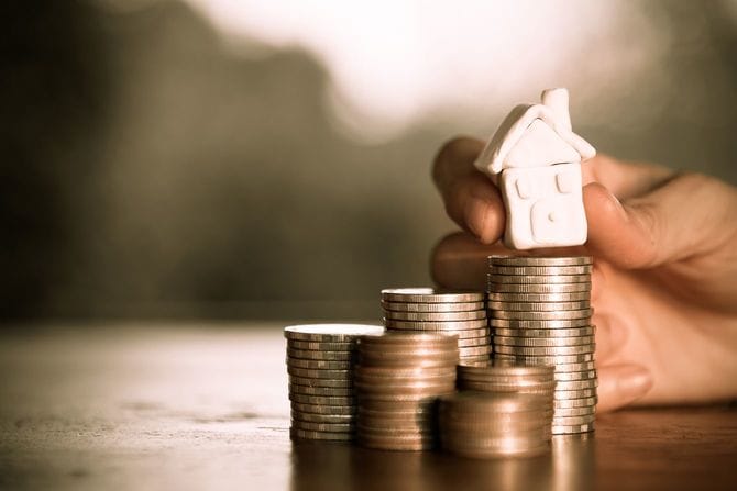 積み上げたコインの上に、粘土で作ったミニチュアハウスを載せようとしている手元