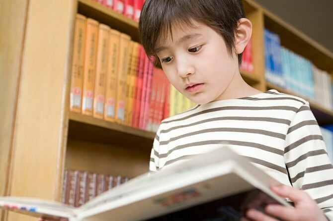 図書館の書棚の前で本を広げて読み始めている少年