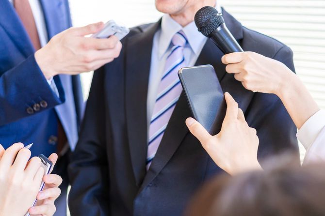 レコーダーを手にした記者たちに囲まれているスーツ姿の男性