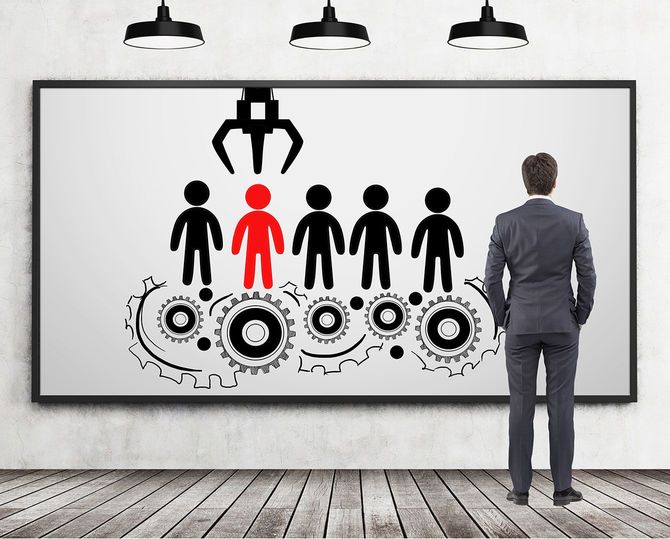 歯車の上に並んだ人型が描かれた絵を見ている男性