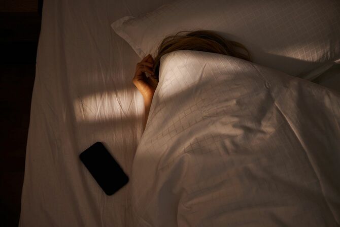 ベッドの上のスマホと、頭まで布団をかぶって寝ている人