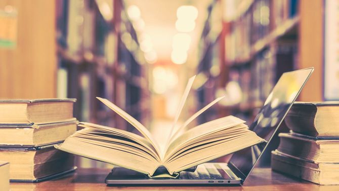 図書館の机の上に、開いて置かれた書籍とノートパソコン