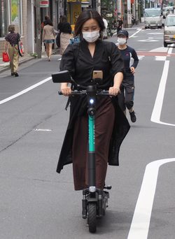 コートを風になびかせながらスマホを注視して走行する電動キックボード