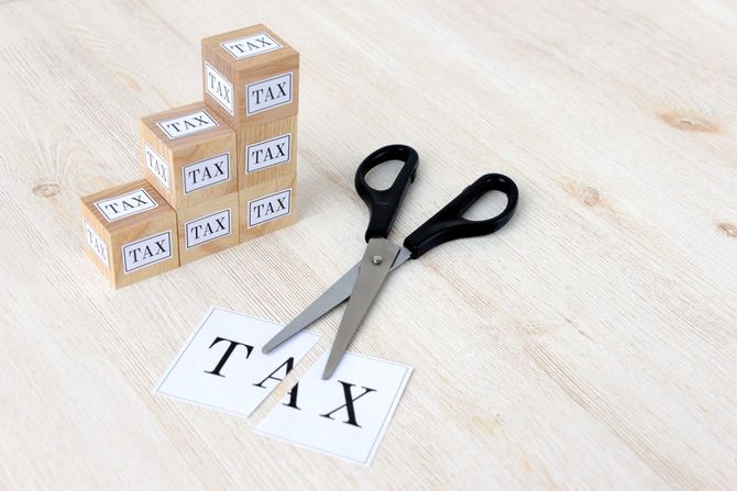 積み上げられた「TAX」と書かれた木製ブロックの横で、「TAX」と書かれたカードがハサミで裁断されている