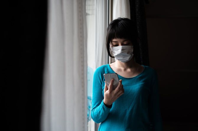 窓にスマートフォンを操作する外科マスクを身に着けているアジアの女性